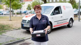 Mitarbeiterin des Malteser Menueservice trägt eine Warmhaltebox und lächelt in die Kamera.