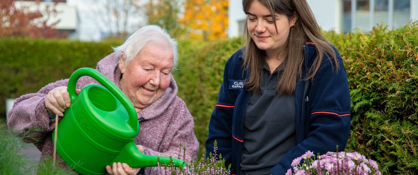 Ältere Frau gießt mit Malteser Helferin im Garten Blumen. 