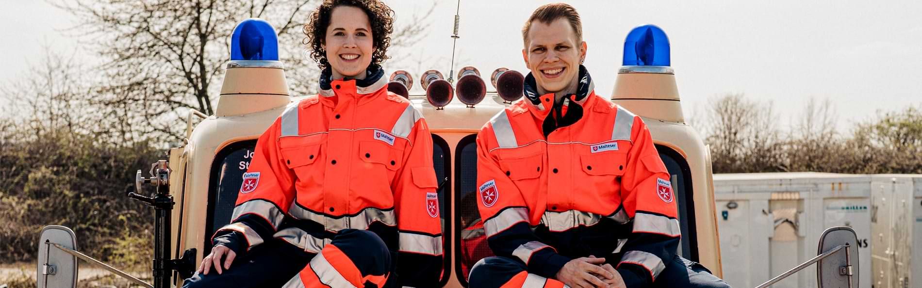 Eine Frau und ein Mann in Einsatzkleidung auf einem Fahrzeug mit Blaulicht.