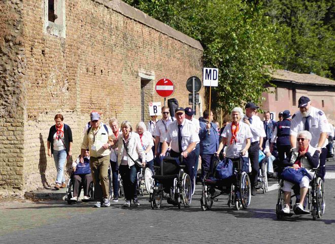 Gruppe von Menschen, darunter mehrere im Rollstuhl, geht eine Straße entlang neben einer hohen Steinmauer mit Verkehrsschildern, darunter ein rotes Einfahrtverbotsschild.