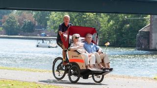 Zwei Senioren werden mit einer Malteser Rikscha an einem Fluss entlang gefahren.