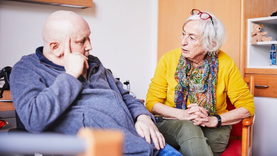 Eine ältere Frau sitzt neben einem Mann mit Glatze, der im Rollstuhl sitzt.