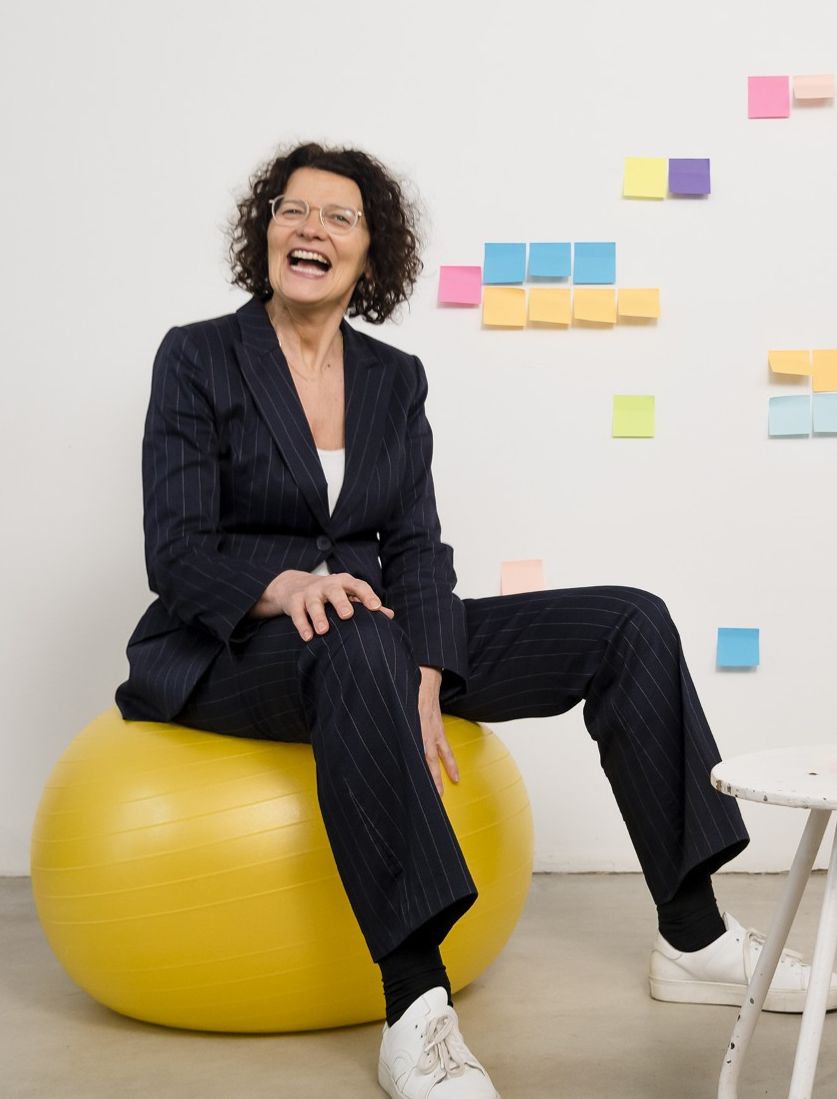 Eine Frau sitzt auf einem gelben Gymnastikball in einem freundlichen Coaching‑Umfeld. Sie lächelt und vermittelt Stabilität und Zuversicht.