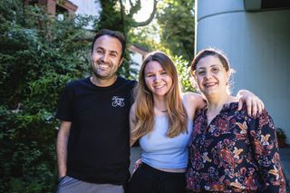Zwei Frauen und ein Mann stehen nebeneinander, umarmen sich und lächeln in die Kamera. 