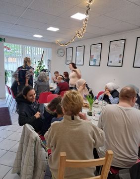 Menschen verschiedener Altersgruppen sitzen an mehreren Tischen in einem hellen Raum und kommen bei Kaffee, Kuchen und Spielen ins Gespräch. Der Treffpunkt ist festlich dekoriert, im Hintergrund sind Ehrenamtliche der Malteser zu sehen.