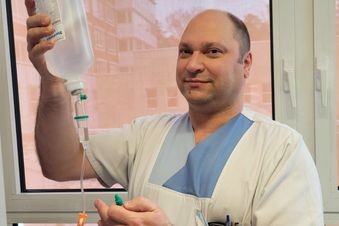 Pflegekraft in weißer Kleidung hält Infusionsflasche hoch und prüft den Schlauch vor einem Fenster mit Tageslicht.