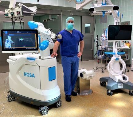 Person steht neben dem Operationsroboter ROSA, im Raum befinden sich Bildschirme und weitere Medizintechnik.
