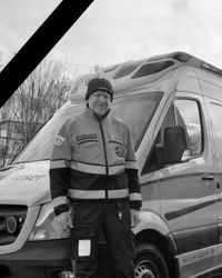 Eine Person in Einsatzkleidung der Malteser steht vor einem Einsatzfahrzeug. Das Bild ist in Schwarz-Weiß gehalten, oben links befindet sich ein Trauerband. Es handelt sich um René Kluge.