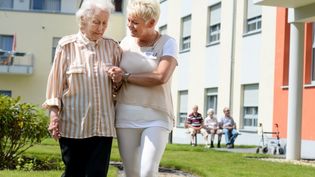 Malteser-Mitarbeiterin führt eine ältere Dame vor einem Pflegeheim