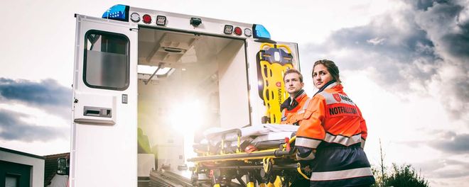 Ein Mann und eine Frau stehen an einer Trage hinter einem Rettungswagen.