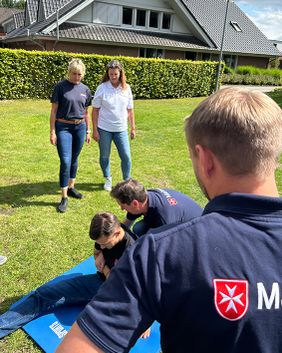 Drei Personen stehen auf dem Rasen in der Sonne. Auf der einer Matte wird von weiteren Personen eine Erst-Hilfe-Übung vorgemacht.