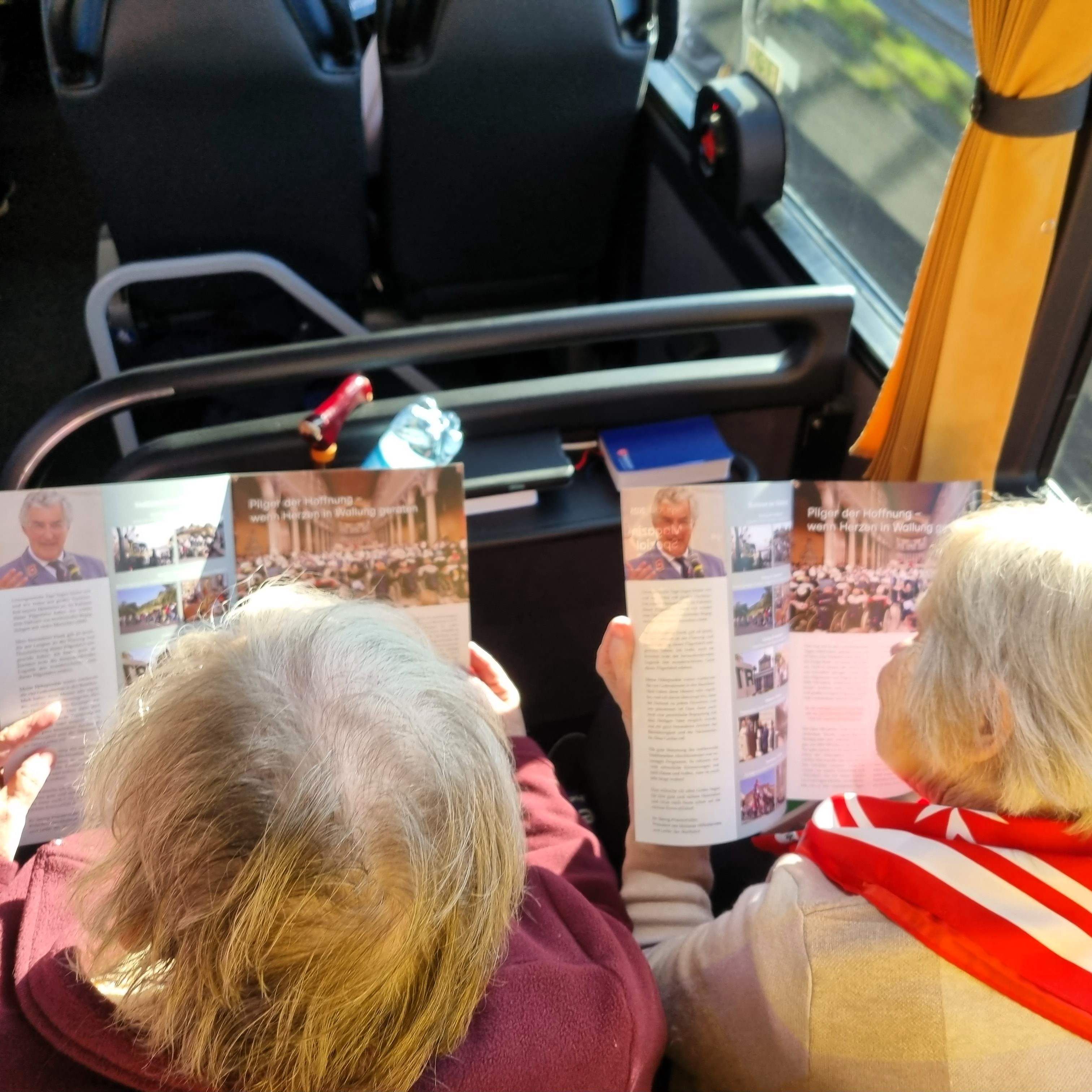 Zwei ältere Personen von hinten, die in einem Fahrzeug sitzen und jeweils eine mehrseitige Broschüre lesen.