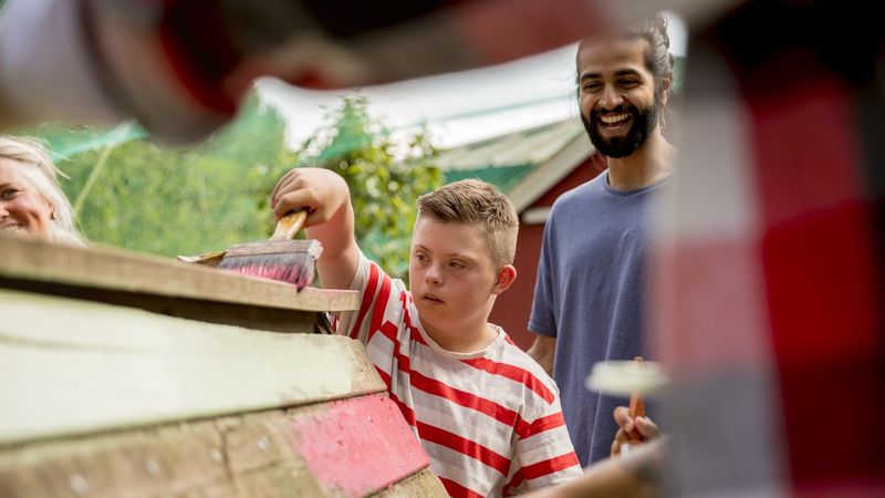 Inklusion in Deutschland: Junge streicht mit einem Betreuer eine Holzhütte an
