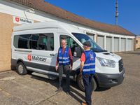 Zwei Ehrenamtliche der Malteser präsentieren die neuen, blauen Warnwesten mit Logo des Mobilen Einkaufswagens vor einem weißen Transporter