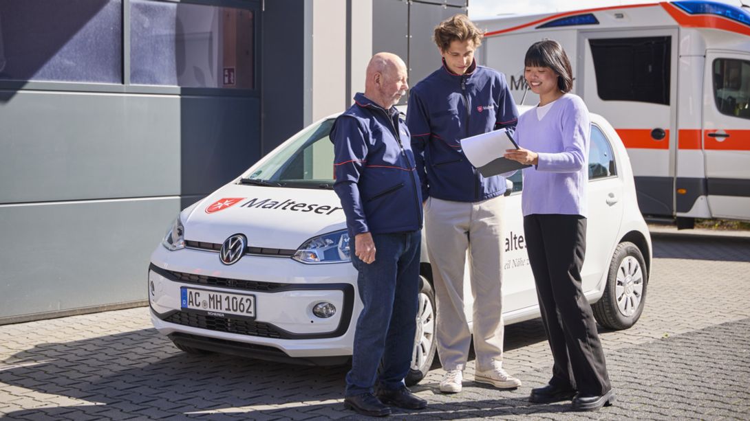 Ein Malteser Hausnotrufmitarbeiter steht vor seinem Auto und spricht mit einer Kollegin und einem Kollegen über einen Dienstplan.