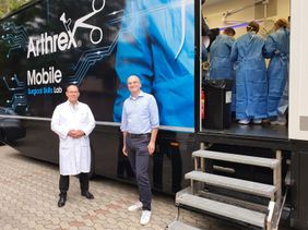 Zwei Männer stehen vor einem mobilen Trainingslabor, in dem Personen in OP-Kleidung arbeiten.