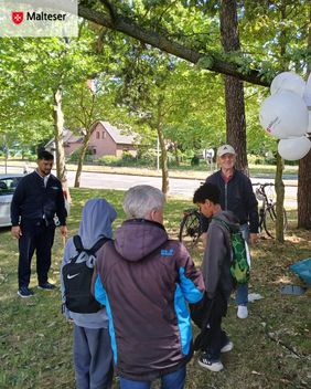 Mehrere Besucherinnen und Besucher stehen auf einer Grünfläche unter schattigen Bäumen am Malteser-Stand und nehmen an einem Spielangebot teil. Rechts im Bild sind weiße Malteser-Luftballons zu sehen, im Hintergrund eine Straße sowie Fahrräder.