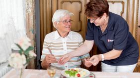 Eine Malteser Mitarbeiterin serviert einer älteren Dame ein Menü.