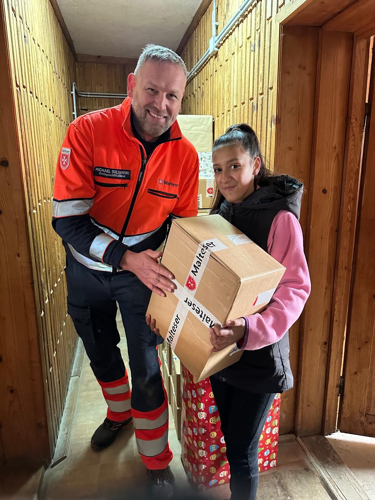 Kind mit einem Paket und Mann in Malteser Kleidung in einem Hausflur