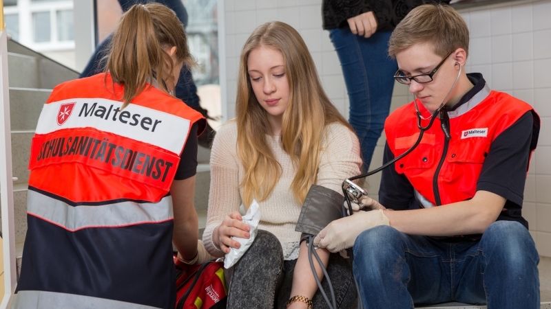 Ein Junge und ein Mädchen des Schulsanitätsdienstes der Malteser versorgen das Knie einer Schülerin. 