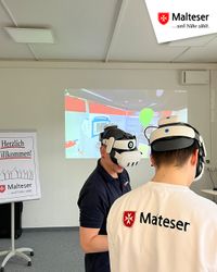 Zwei Personen tragen Virtual-Reality-Headsets und stehen in einem Schulungsraum der Malteser. Auf der Wand hinter ihnen ist eine virtuelle Szene eines Rettungswagens projiziert. Links im Bild steht ein Aufsteller mit der Aufschrift „Herzlich willkommen!“ und dem Malteser-Logo. In der Ecke oben rechts ist ebenfalls das Malteser-Logo eingeblendet.