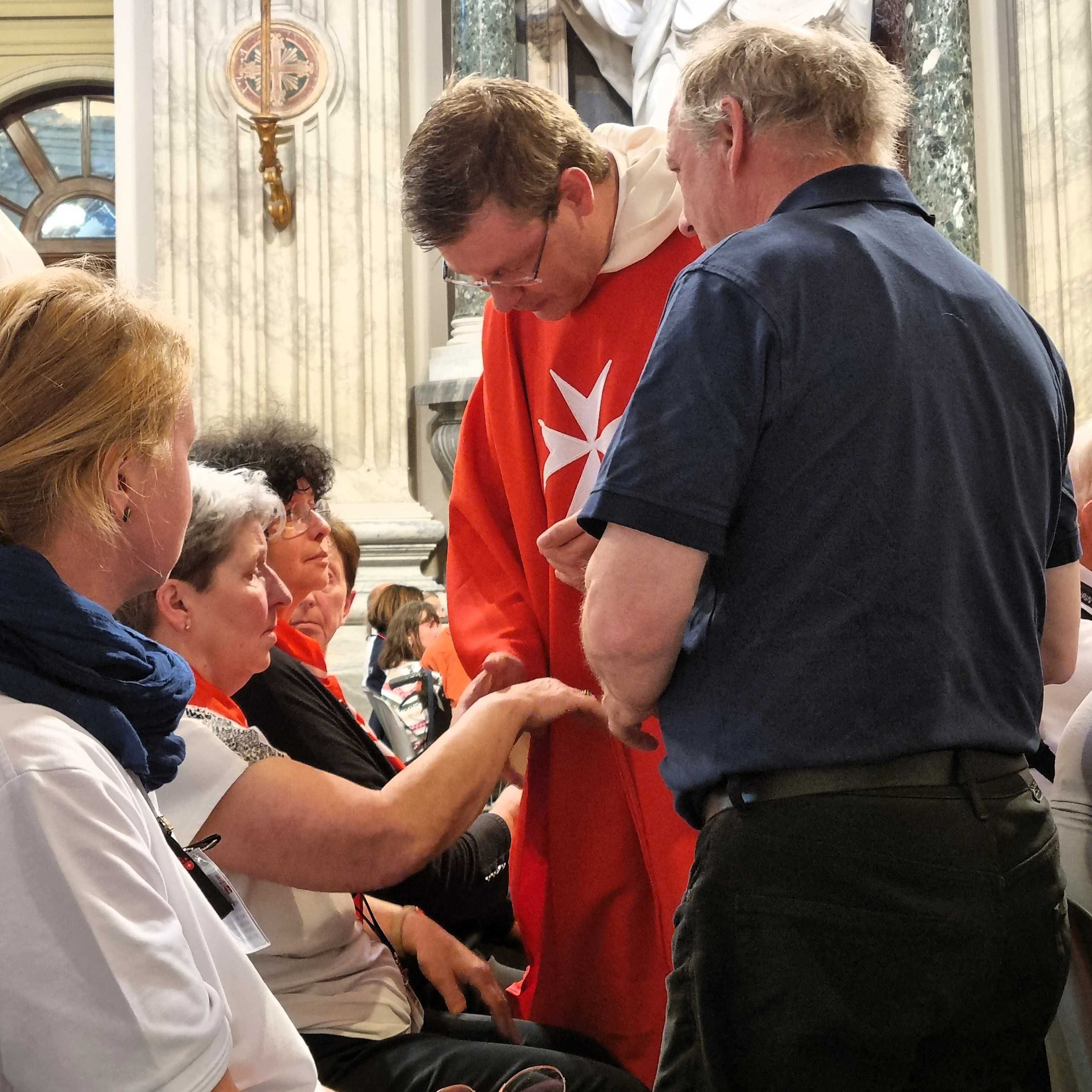 Person in rotem Gewand mit weißem Malteserkreuz steht in Kirche, hält Hand einer sitzenden Person, weitere Personen im Hintergrund sichtbar.