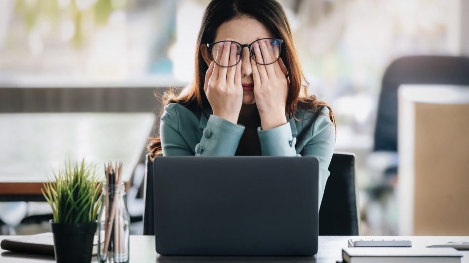 Eine Frau, die an einem Schreibtisch vor einem Laptop sitzt, reibt sich erschöpft die Augen.