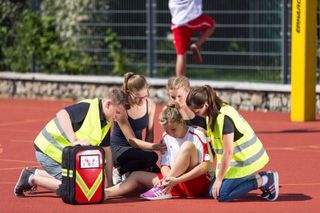 Schülerinnen und Schüler in gelben Warnwesten knien um ein am Bein verletztes Mädchen auf einem roten Sportplatz