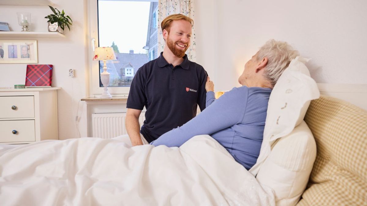 Ein Mitarbeiter der ambulanten Pflege der Malteser mit einer Patientin, die im Bett liegt.
