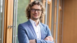 Businessporträt einer Person mit lockigem Haar und Brille im Anzug vor großen Fenstern.