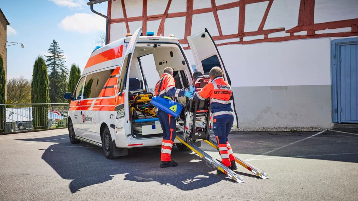 Malteser in Einsatzkleidung helfen einem gehbehindertem Mann in in ein Fahrzeug.