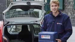 Ein junger Mann in Malteser-Kleidung steht mit einer Isolierbox in den Händen vor der geöffneten Heckklappe eines Autos.