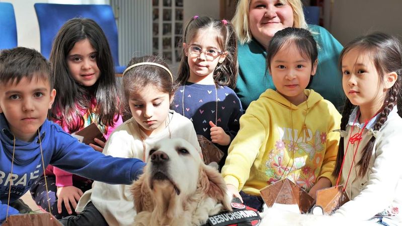 Sechs Kinder und eine lächelnde Frau mit einem Hund, der ein Geschirr mit der Aufschrift „Besuchshund“ trägt.