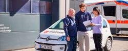 Frau mit Klemmbrett steht neben zwei Männern. Alle stehen vor einem Auto.
