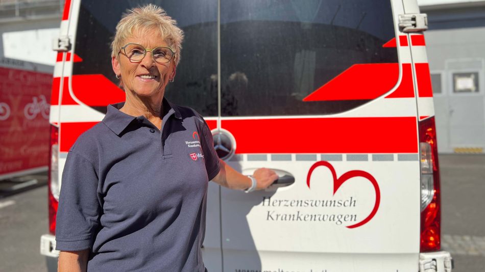 Eine Frau steht vor einem Malteser Herzenswunsch Krankenwagen und hält die Hecktür fest.