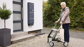 Eine ältere Frau mit Rollator und Mobilnotrufuhr am Handgelenk steht vor einem Hauseingang.