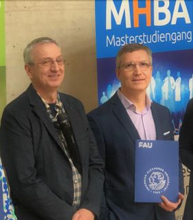 Zwei Männer im Anzug stehen vor einem Banner, einer hält eine Mappe der FAU Universität.