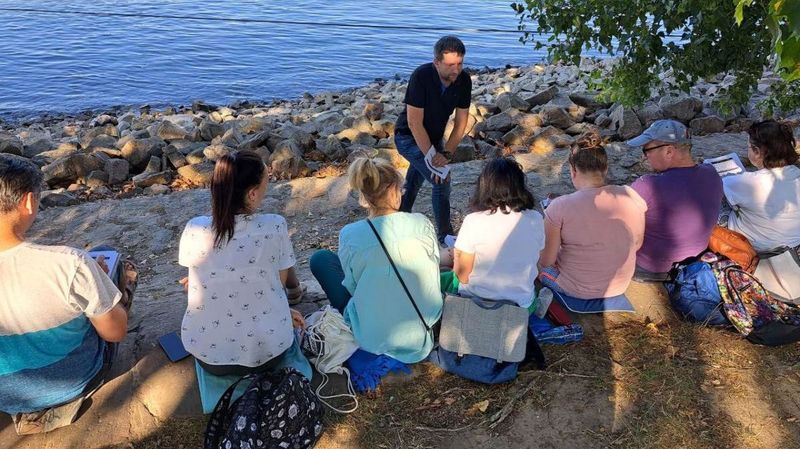 Sieben sitzende Menschen an einem Fluss und ein Mann, der vor ihnen steht.