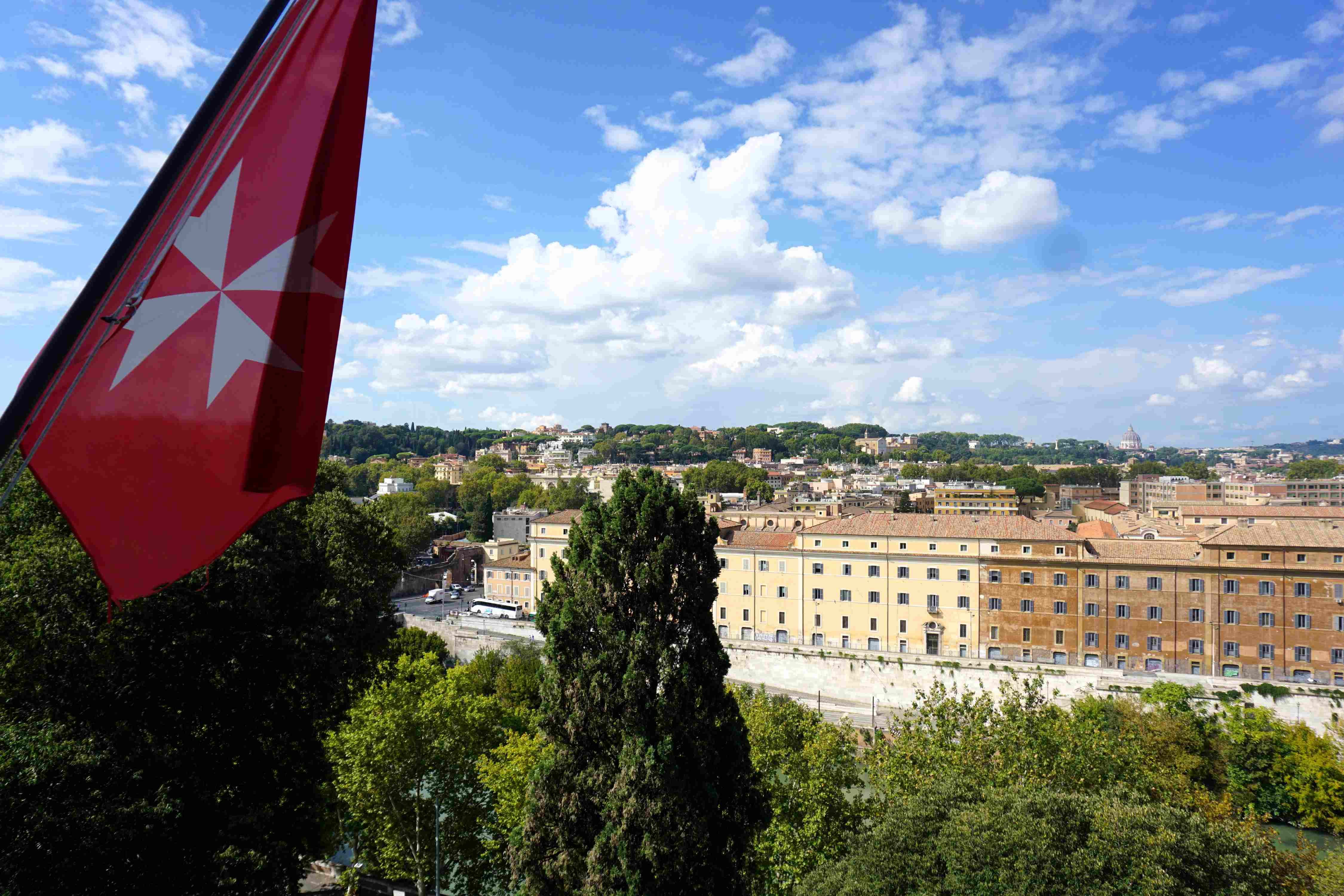 Rotes Flaggenbanner mit weißem Malteserkreuz vor Stadtansicht mit Bäumen und Gebäuden unter blauem Himmel mit Wolken.