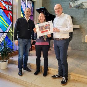 Drei Personen in einer Kapelle mit bunten Fenstern halten einen Spendenscheck.
