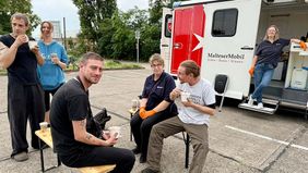 Menschen sitzen auf klappbaren Bänken. Im Hintergrund ist das MalteserMobil zu sehen.