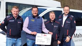 Gruppenfoto der an der Übergabe des symbolischen Spendenschecks beteiligten Personen vor einem Fahrzeug der Malteser