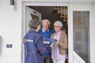 Älteres Ehepaar steht an der Haustür und bekommt von einem Malteser eine Box überreicht