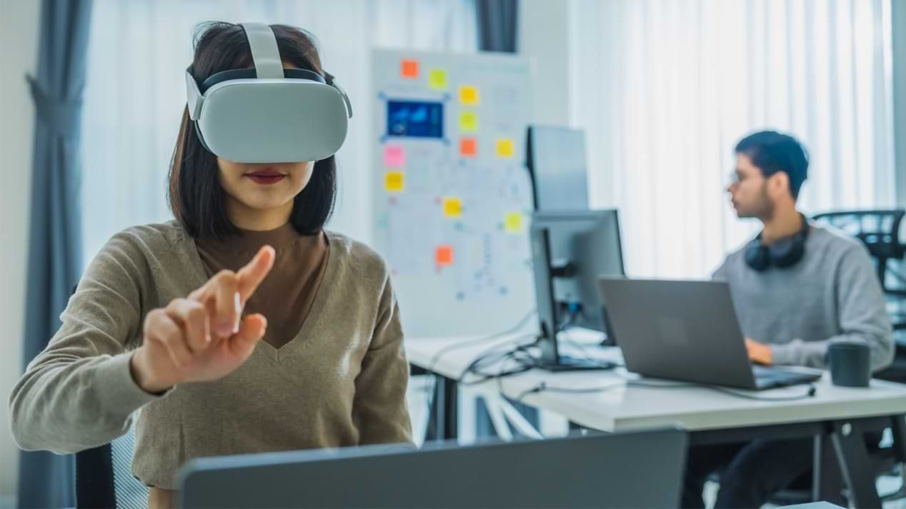 Eine Frau mit VR-Brille im Vordergrund, dahinter ein Mann mit Laptop und einem Whiteboard.