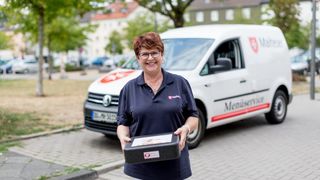 Mitarbeiterin des Malteser Menueservice trägt eine Warmhaltebox und lächelt in die Kamera.