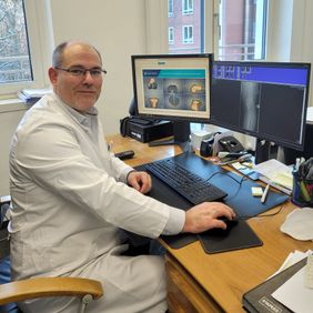Arzt im weißen Kittel sitzt am Schreibtisch und betrachtet Röntgen- und 3D-Bilder auf zwei Monitoren.