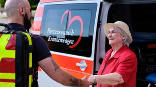 Mann mit Rucksack und tätowiertem Arm schüttelt Hand einer älteren Frau mit Strohhut vor einem orange-weißen Krankenwagen mit Aufschrift 'Herzenswunsch Krankenwagen'.