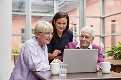 Eine ältere Frau und ein älterer Mann sitzen am Laptop. Zwischen beiden steht eine junge Frau der Malteser, die ihnen etwas erklärt. (Foto: Malteser) 