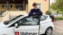 Ein junger Mann in Malteser-Kleidung steht in der Tür eines Malteser-Autos.