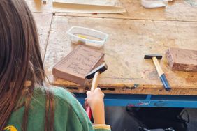 Eine Person sitzt an einer Werkbank und bearbeitet mit Werkzeugen eine Steinplatte. Auf dem Tisch liegen weitere Materialien und Arbeitsgeräte.
